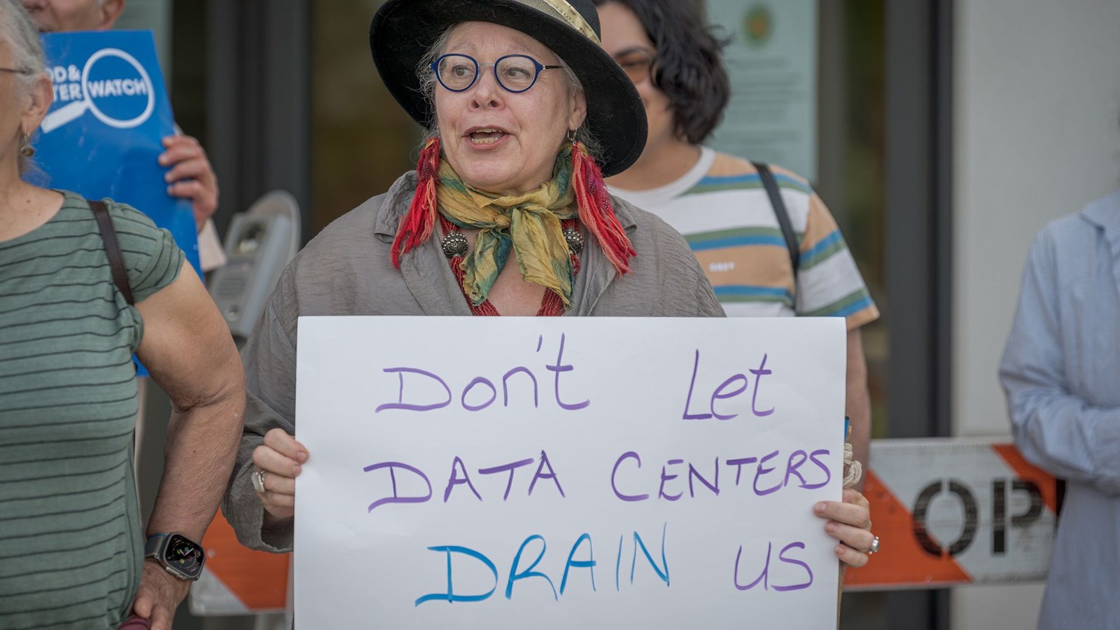 A woman wearing a dark hat and round glasses holds a protest sign.