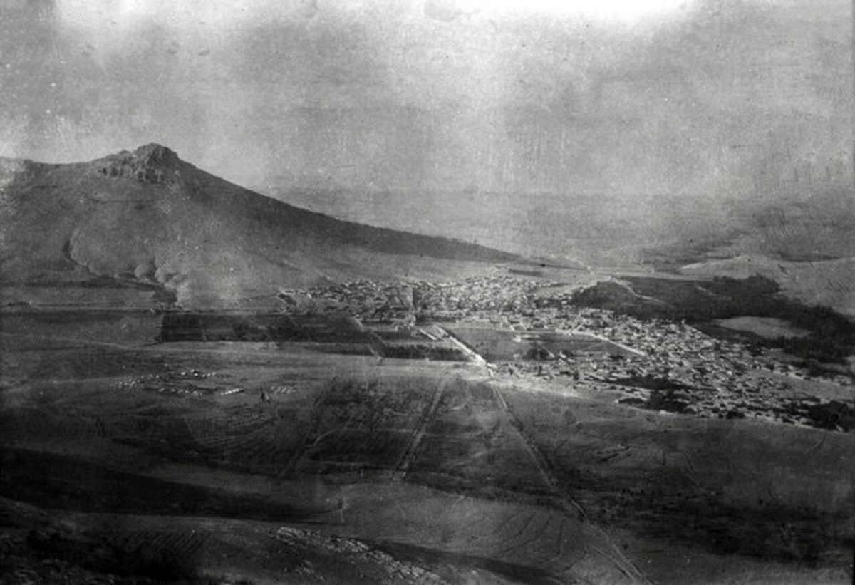 A hazy black and white view of the Persian town of Bijar spread across a valley floor beneath a bare hill, photographed from high ground