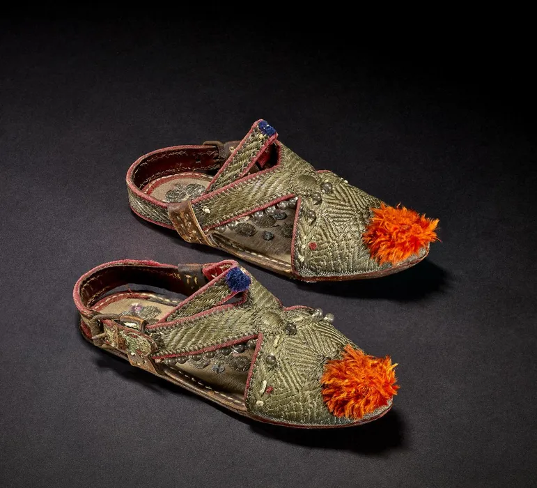 Pair of children's (boy's?) closed-toe leather and cotton sandals. Decorated with gilt-metal wrapped thread embroidery, glass beads and orange and blue silk pom-poms. With a metal buckle closure on one side and red cotton detailing along the edges. - Bilinmiyor